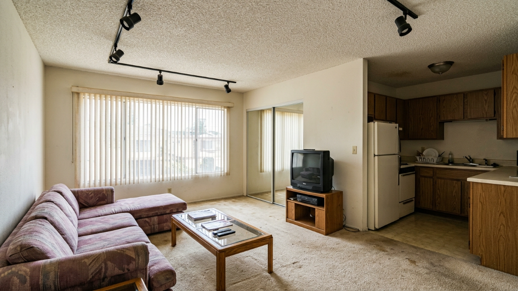 Bare 90s apartment with vertical blinds, mauve plaid sofa, glass coffee table, oak kitchen, and CRT TV